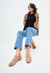Beige suède loafers met een gestikte accent, casual blauwe denim flare broeken en een gestructureerde zwart-en-tan cardigan. Minimalistische houten kruk.