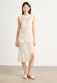 Knit cream-colored dress with a fitted design, sleeveless cut, side slit, and floral patterns integrated into the fabric texture.