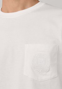 Camiseta blanca de cuello redondo con un bolsillo en el pecho que presenta un emblema en relieve con "1950" y detalles de corona de laurel.