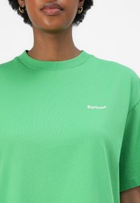 Zelené tričko s krátkým rukávem vyrobené z měkké bavlny, s vroubkovaným kulatým výstřihem a malým bílým logem "Barbour" na levé straně hrudi.