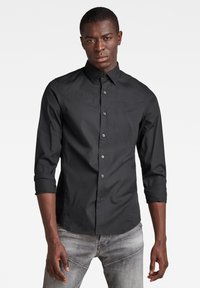 Camicia nera con bottoni, maniche lunghe, caratterizzata da un colletto classico e un piccolo logo. Realizzata in un tessuto morbido con un design aderente.