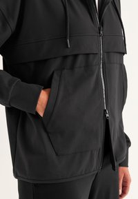 Zwarte ritssluitingjack met hoge kraag, voorzakken, een verstelbare capuchon en een gladde, gestructureerde stof. Opvallende stiknaaddetails over de borst.