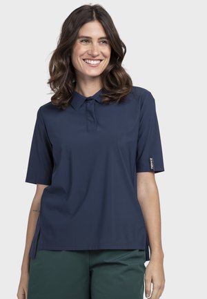 Lächelnde Frau mit welligem braunem Haar, die ein marineblaues Poloshirt mit kurzen Ärmeln und dunkelgrüne Hosen vor einem einfarbigen hellen Hintergrund trägt.