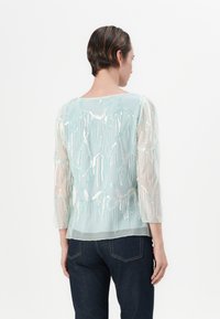 Blusa traslucida blu chiaro con maniche a 3/4, caratterizzata da decorazioni di perline iridescenti e fili, che creano un design testurizzato sulla schiena.