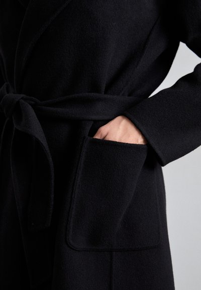 Zwarte wikkelcoat van zachte, gestructureerde stof. Heeft een strikceintuur en een grote opgestikte zak aan de linkerkant.