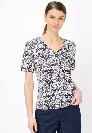 Frau trägt ein kurzärmeliges V-Ausschnitt-Oberteil mit schwarzem und weißem Blattmuster, kombiniert mit dunkelblauer Hose, steht vor weißem Hintergrund.