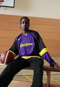 Violeta un melna basketbola krekls ar dzelteniem akcentiem un tekstu "LakeShow", ar tumšām biksēm; tur basketbola bumbu. Koka fons.