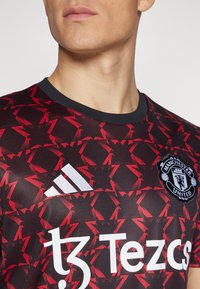 Mustan ja punaisen värinen Manchester Unitedin pelipaita, jossa on geometrinen kuosi. Adidas-logo ja sponsorin "Tezos" printti valkoisena. Lyhyet hihat, pyöreä kaulus.