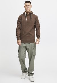 Sudadera marrón con botones de madera en el cuello, combinada con pantalones cargo verde claro. El atuendo presenta un ajuste holgado y detalle de cordón.