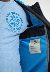 Hellblaues Sweatshirt mit einem Palmbaum-Graphic und Text, darunter eine schwarz-blaue Jacke mit Reißverschluss, Netzfutter und Etikett.