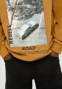 Gelbes Sweatshirt mit einem Grafikdesign eines silbernen Autos aus der Vogelperspektive, auf der Vorderseite in fetter schwarzer Schrift "REBELL" und "ROAD" bedruckt.