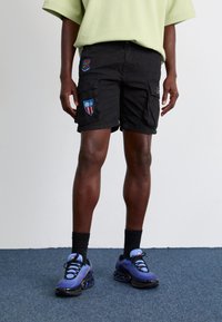 Black cargo shorts with two side pockets, featuring colorful patches on both sides. Worn with blue athletic shoes and black crew socks.