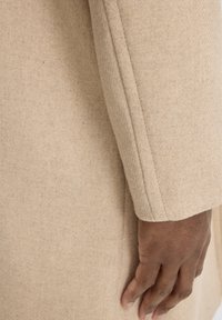 Manica di cappotto beige con tessuto testurizzato, caratterizzata da un taglio dritto e dettagli di cucitura visibili. Mano appoggiata sul tessuto.