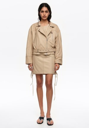 Mujer de cabello oscuro que lleva una chaqueta de cuero beige con cinturón y falda, combinadas con sandalias negras de punta abierta, posando frente a un fondo blanco.