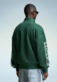 Person iført en mørkegrøn sweatshirt med hvid tekst på ærmerne og grå sweatpants med broderi på lommen, stående foran en ensfarvet baggrund.