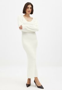 Robe tricotée blanche longue et ajustée avec un décolleté dégagé et des manches bouffantes, complétée par des escarpins noirs à talons pointus. Texture lisse dans l'ensemble.