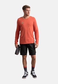 T-shirt arancione a maniche lunghe con design grafico, abbinato a pantaloni corti neri e scarpe alte bianche e nere, mentre tiene una borsa clutch scura.