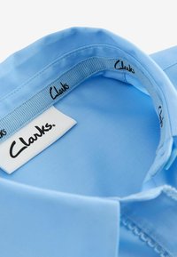 Colletto di camicia azzurro chiaro con una texture liscia. Presenta un'etichetta di marca con "Clarks" in nero. La cucitura è visibile lungo i bordi.