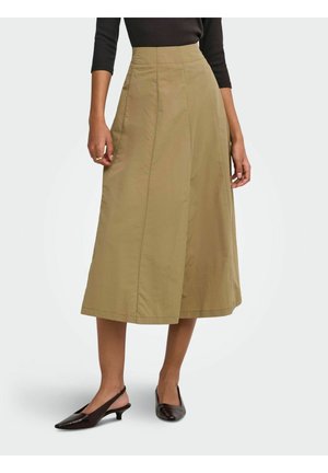 Femme portant une jupe midi beige taille haute, un top noir à manches trois-quarts et des talons slingback noirs pointus, debout sur fond blanc.