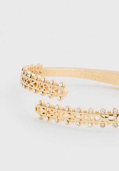 Kuldne metallist käevõru, millel on keerukad väljalõigatud mustrid ja servade ääres selged rhinestone'ide aktsendid, mis rõhutavad õrna ja tekstuuriga viimistlust.