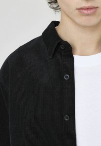 Chemise en velours côtelé noire avec un col pointu et une fermeture à boutons, associée à un T-shirt blanc en dessous. Une texture à côtes verticales est visible.