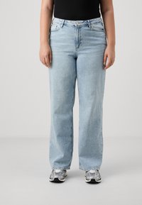 Person wearing light blue straight-leg jeans, black top, and silver-gray sneakers, standing against a plain light gray background.