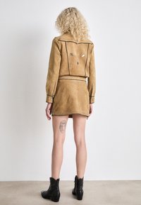 Giacca in suede beige con dettagli in metallo, abbinata a una gonna coordinata con un orlo ondulato e ulteriori borchie, indossata con stivaletti neri.