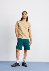 Beige Baumwoll-T-Shirt mit Rundhalsausschnitt, kombiniert mit dunkelgrünen Shorts. Die Shorts haben eine weiche Textur, dazu schwarze Sportschuhe und weiße Socken.