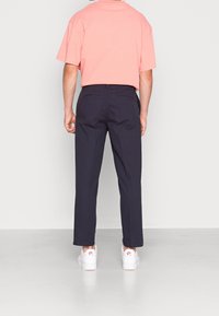 Kurzärmliges, korallfarbenes T-Shirt kombiniert mit marineblauen, geraden Hosen und weißen Sneakers mit roten Akzenten, von hinten betrachtet.