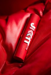 Röd cylindrisk enhet med vit text "JAYIT™" på en texturerad röd tygbakgrund. Har en jämn yta och rundade ändar.