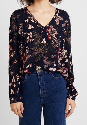 Femme portant un chemisier bleu marine à fleurs, col en V et manches longues, rentré dans un jean bleu taille haute, sur un fond uni.