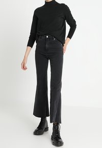 Pull noir à col montant, associé à un jean flare noir taille haute avec des ourlets bruts. Porté avec des bottes noires à lacets.