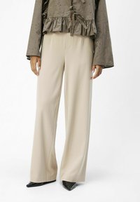 Beige Wide-Leg-Hosen mit glatter Textur, getragen mit einem dunkelgrünen, rüschengeschmückten Oberteil mit Pünktchen. Spitzer schwarzer Absatzschuhe runden das Outfit ab.