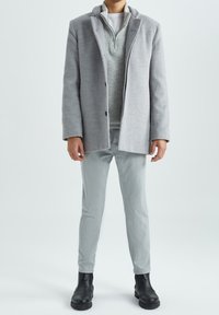 Abrigo gris con cuello en muesca y cierre de un solo botón, superpuesto a un suéter gris claro con cremallera, combinado con pantalones gris claro y botas negras.