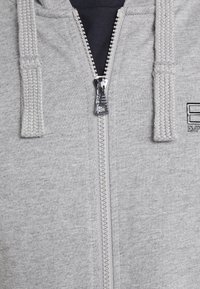 Sudadera con cremallera en gris, con cordones trenzados y tejido texturizado. Presenta una cremallera plateada y un parche con el logo en la parte frontal.