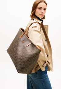 Γυναίκα που φοράει ένα μπεζ παλτό τύπου trench και μπλε τζιν, κρατώντας μια μεγάλη καφέ τσάντα ώμου με σχέδια, στραμμένη ελαφρώς προς τα πλάγια.