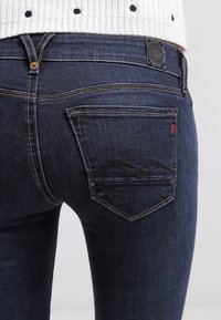 Vaqueros de mezclilla azul oscuro con bolsillos traseros con detalle de costura y un pequeño acento rojo. La tela tiene una textura suave.