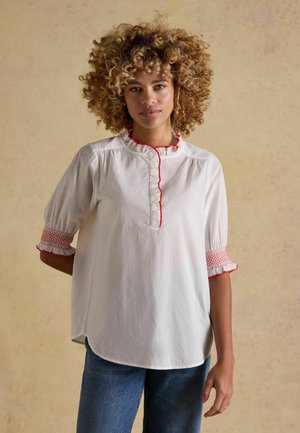 Žena s kučeravými vlasmi oblečená v bielej blúzke s červeným lemom na golieri a rukávoch, stojaca pred béžovým textúrovaným pozadím.