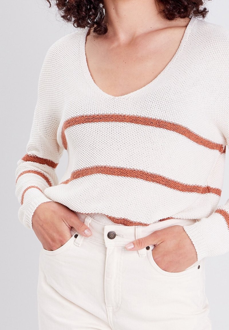Femme portant un pull en maille blanc avec des rayures horizontales couleur rouille et un jean blanc, les mains partiellement dans les poches avant.