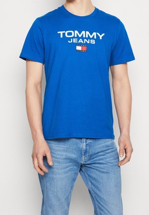 Mand iført en klar blå Tommy Jeans t-shirt og lyseblå denimjeans, stående med armene afslappet ned langs siderne.