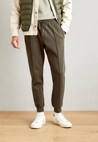 Mörk olivgröna joggingbyxor med dragsko i midjan, tapered fit och sidosemdetaljer, tillsammans med vita sneakers på ett trägolv.