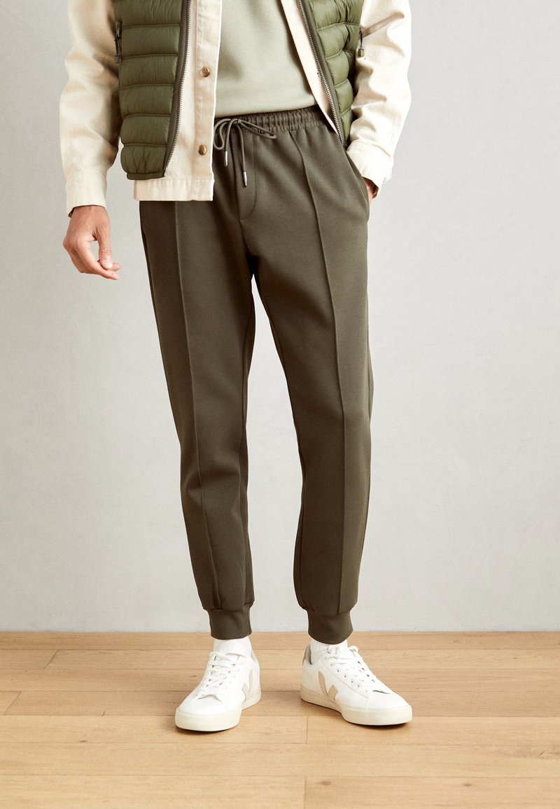 Mörk olivgröna joggingbyxor med dragsko i midjan, tapered fit och sidosemdetaljer, tillsammans med vita sneakers på ett trägolv.