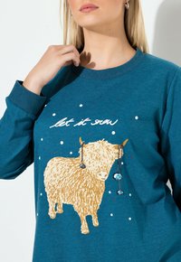 Maglietta a maniche lunghe color turchese con una grafica di una mucca delle Highlands dorata, decorata con punti bianchi simili alla neve e la scritta "let it snow" in bianco.