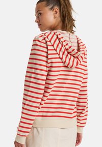 Sudadera a rayas en colores rojo y crema, con capucha ajustable, puños acanalados y un corte relajado. Tejido suave con una textura lisa.