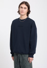 Jeune homme aux cheveux bruns mi-longs, portant un sweat-shirt Volcom bleu foncé et un jean gris sur un fond blanc uni.