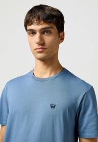 Blauwe t-shirt met ronde hals van zacht materiaal. Bevat een klein marineblauw logo op de borst. Gladde textuur met korte mouwen.
