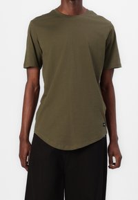 Camiseta de algodón verde oliva con cuello redondo, mangas cortas y dobladillo curvado. Presenta una pequeña etiqueta en la costura lateral.
