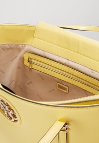 Żółta syntetyczna torebka o gładkiej fakturze, z zamykanym na zamek wewnętrznym schowkiem i logo na przodzie, ozdobiona jasną podszewką.