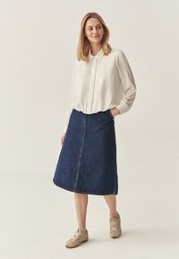 Biała koszula na guziki z kołnierzykiem, zestawiona z ciemną dżinsową spódnicą o długości do kolan w kształcie litery A oraz beżowymi trampkami. Prosty i codzienny fason.