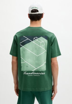 Čovjek koji nosi zelenu majicu i bijelu kapu, na stražnjoj strani majice je apstraktni grafički prikaz nogometnog igrališta s tekstom "Scandinavian Football Community".